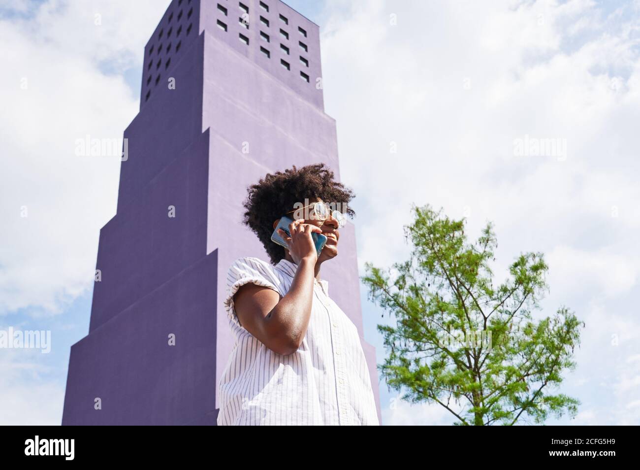 Vue d'en dessous de côté de la femme afro-américaine mince robe blanche et bottes regardant loin et parlant au téléphone tout en se tenant seul contre le bâtiment violet de plusieurs étages dans la ville à jour d'été ensoleillé Banque D'Images