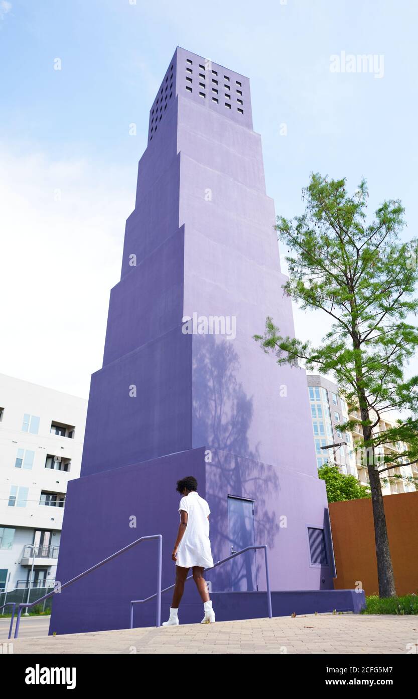 De l'arrière-plan de la femme afro-américaine mince en robe blanche et des bottes marchant seul contre le bâtiment violet de plusieurs étages dans la ville en jour ensoleillé d'été Banque D'Images