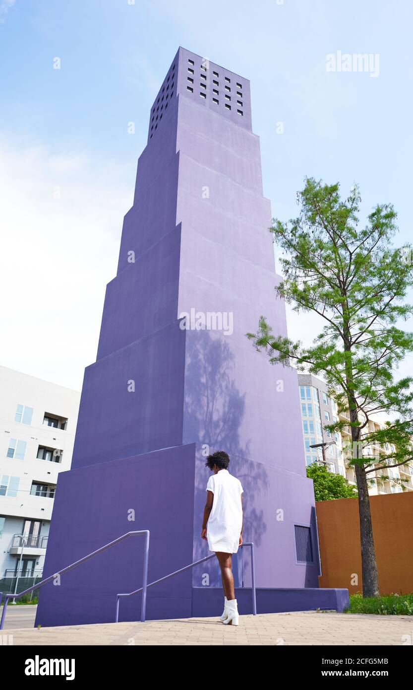 De l'arrière-plan de la femme afro-américaine mince en robe blanche et des bottes marchant seul contre le bâtiment violet de plusieurs étages dans la ville en jour ensoleillé d'été Banque D'Images