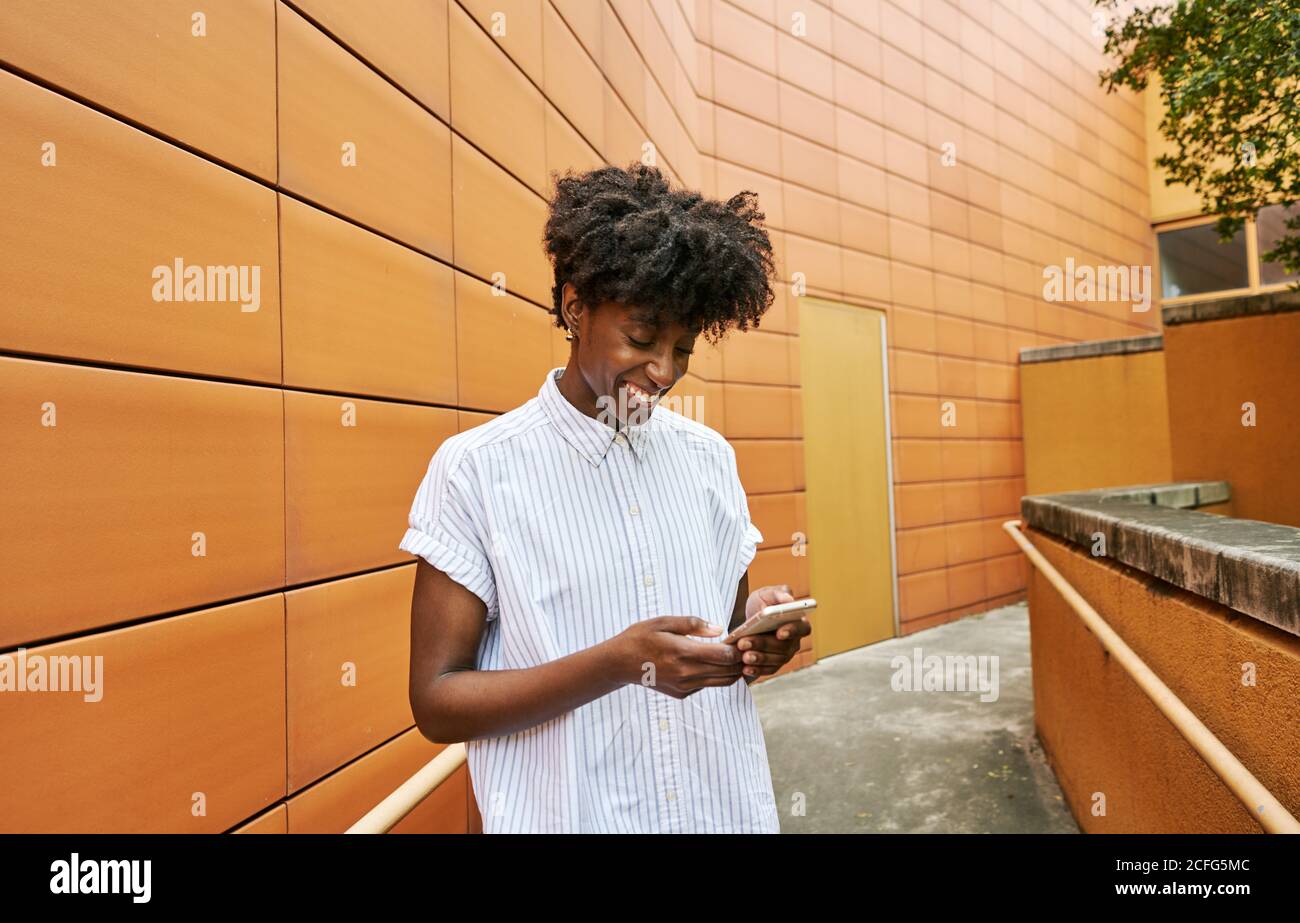 Adulte Afro-américaine femme en blanc décontracté chemise souriant pendant messagerie sur smartphone debout dans la rue, avec un carrelage orange vif façade de bâtiment contemporain dans le centre-ville Banque D'Images