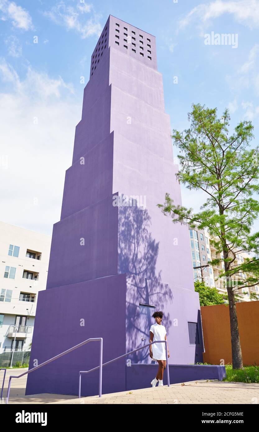 De dessous de vue de la femme afro-américaine mince en robe blanche et des bottes marchant seul contre le bâtiment violet de plusieurs étages dans la ville dans la journée ensoleillée d'été Banque D'Images