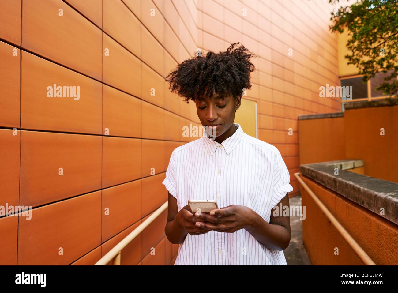 Adulte Afro-américaine femme en blanc décontracté message sur le maillot smartphone situé dans la rue, face à la façade en céramique orange vif de bâtiment contemporain au centre-ville Banque D'Images