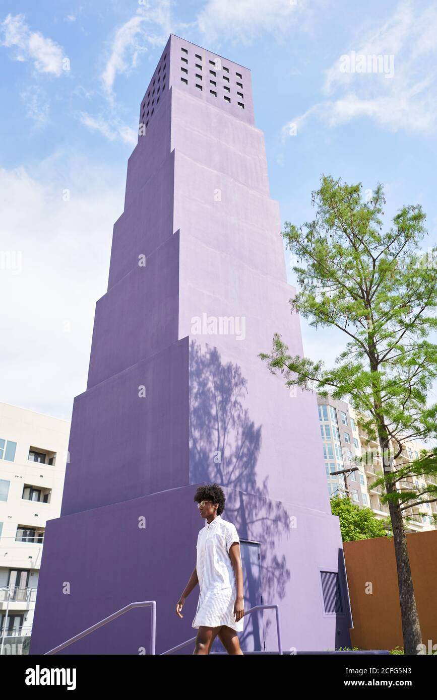 De dessous de vue de la femme afro-américaine mince en robe blanche et des bottes marchant seul contre le bâtiment violet de plusieurs étages dans la ville dans la journée ensoleillée d'été Banque D'Images