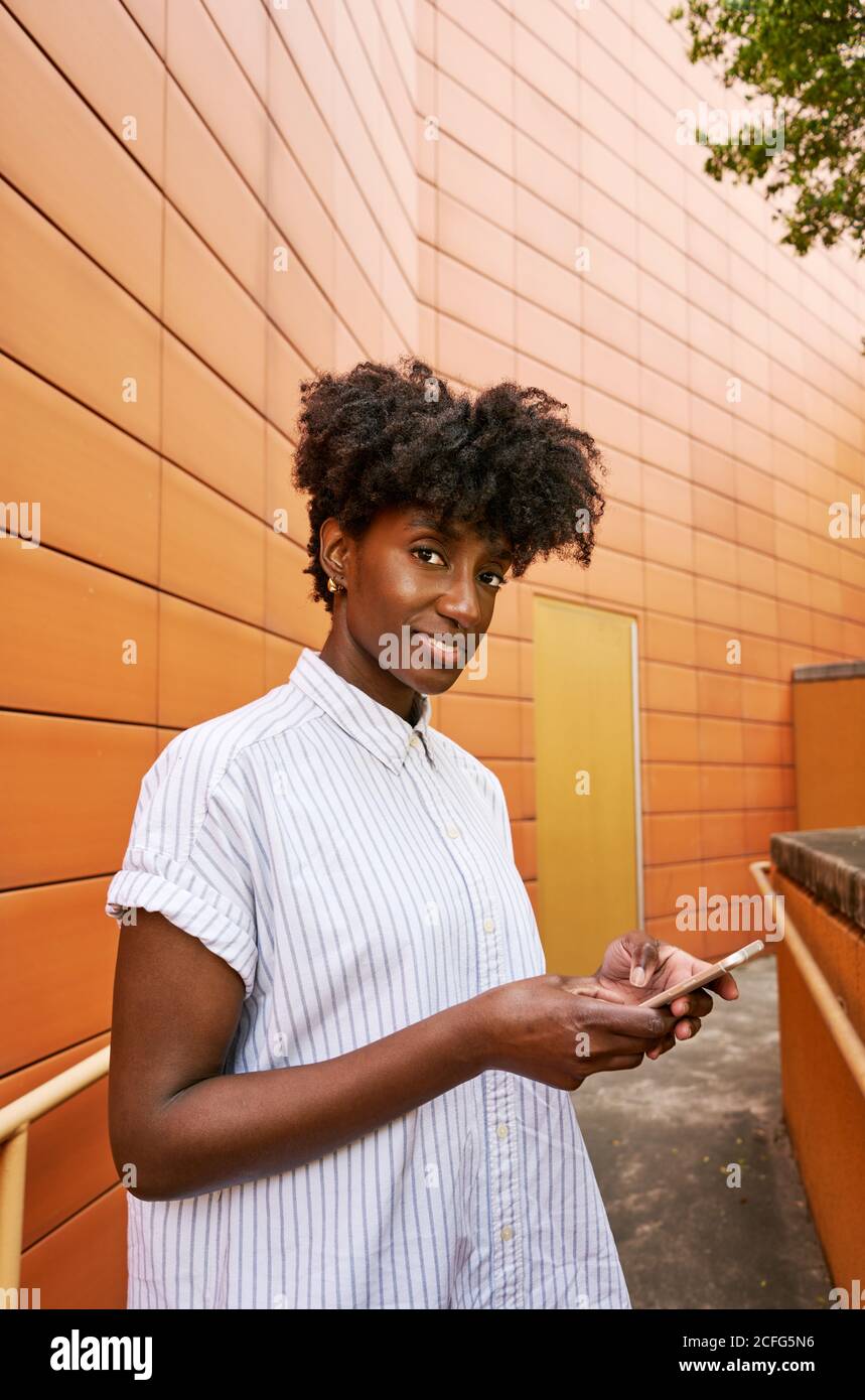 Adulte Afro-américaine femme en blanc décontracté chemise souriant un appareil photo tout en envoyant des messages sur votre smartphone, dans la rue, contre les couleurs vives façade de carrelage orange d'un bâtiment contemporain dans le centre-ville Banque D'Images