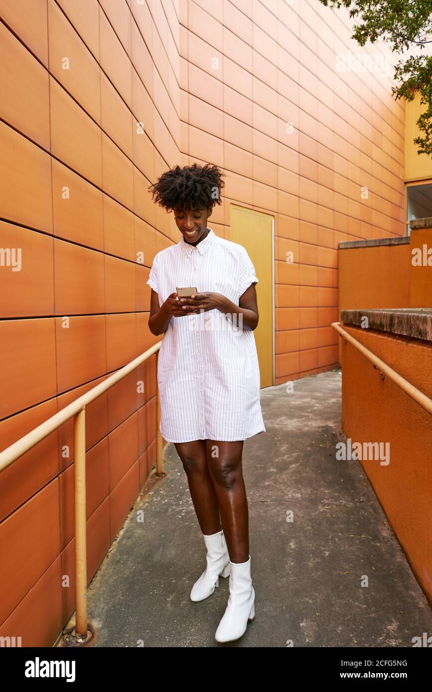 Adulte Afro-américaine femme en blanc décontracté message sur le maillot smartphone situé dans la rue, face à la façade en céramique orange vif de bâtiment contemporain au centre-ville Banque D'Images