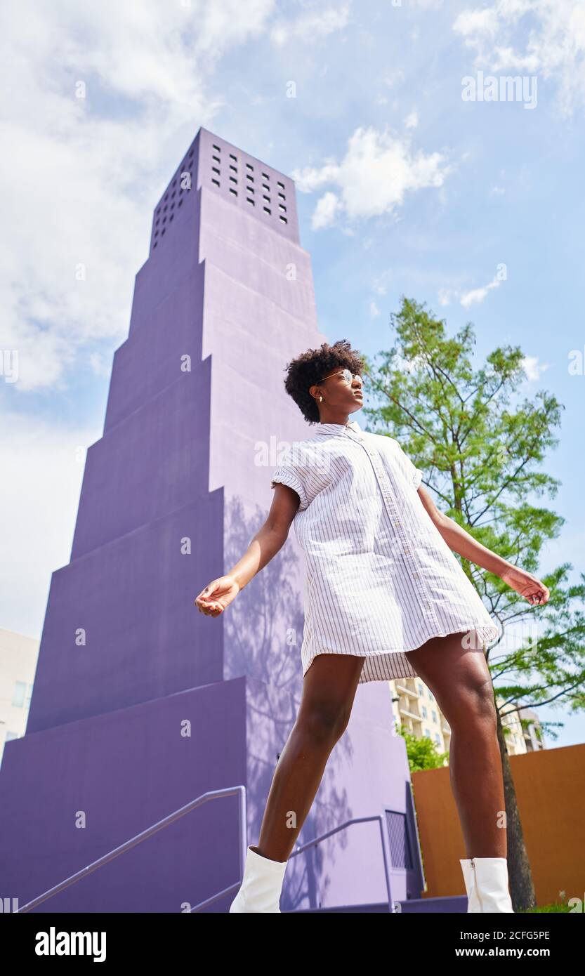 De dessous de vue de femme afro-américaine mince en robe blanche et des bottes posant seul contre le bâtiment violet de plusieurs étages dans la ville en jour ensoleillé d'été Banque D'Images