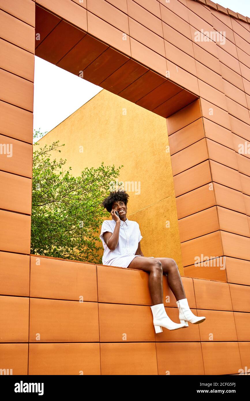 Angle bas de la jeune femme afro-américaine dans le blanc tendance habillez-vous en souriant et en téléphonant dans la rue installé dans un bâtiment contemporain aux murs orange et jaune vif murs Banque D'Images