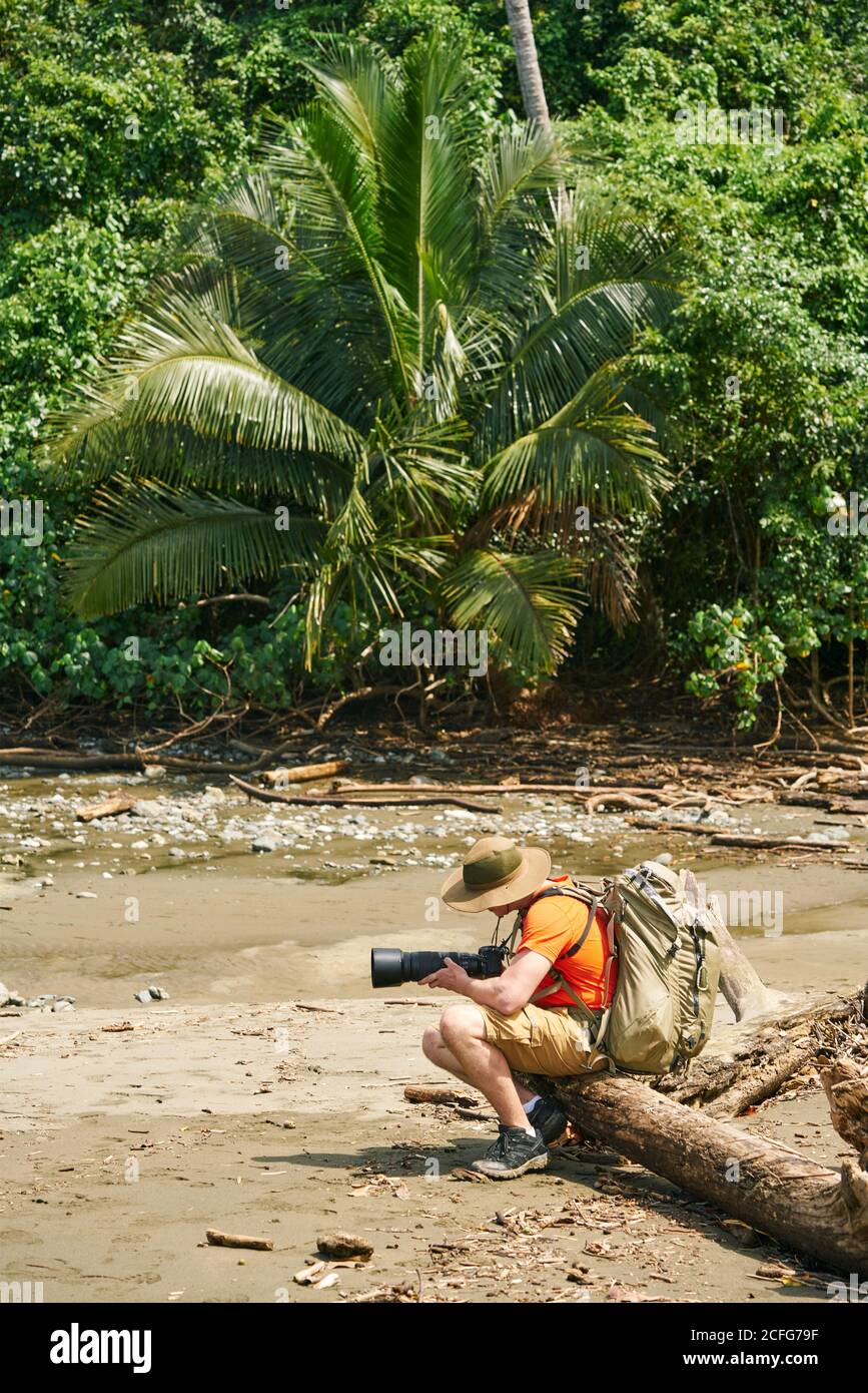 Vue latérale d'un touriste anonyme avec sac à dos pour prendre des photos de paysage avec un appareil photo professionnel assis dans un arbre branche sur terre au Costa Rica Banque D'Images