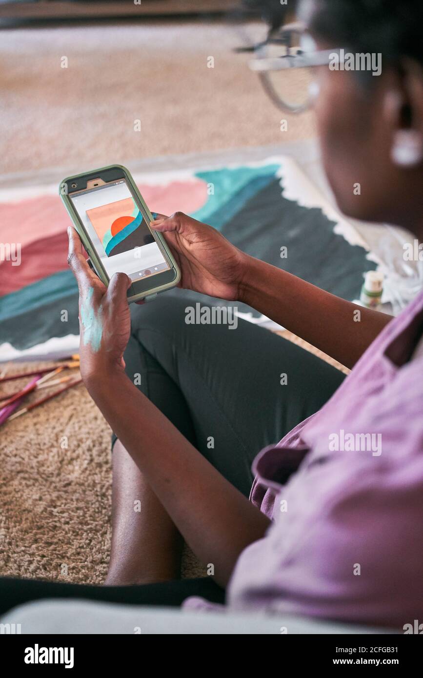 Vue latérale d'une femme afro-américaine en utilisant l'application avec échantillon d'image sur smartphone tout en apprenant à peindre à la maison pendant temps libre Banque D'Images
