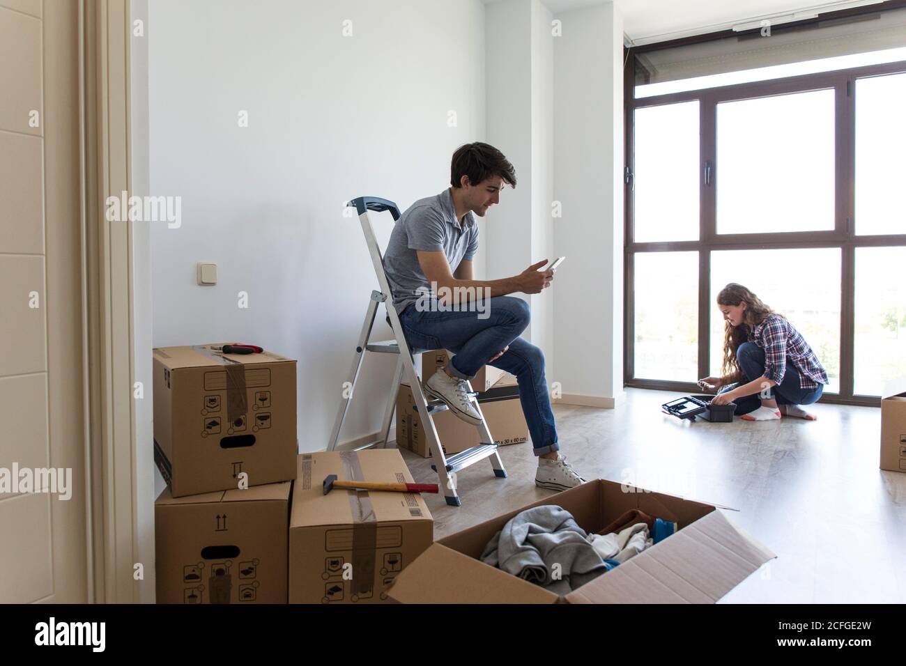 Couple d'hommes assis sur une échelle à l'aide d'un téléphone portable et femme prenant les outils de la boîte dans l'appartement vide avec la boîte boîtes Banque D'Images