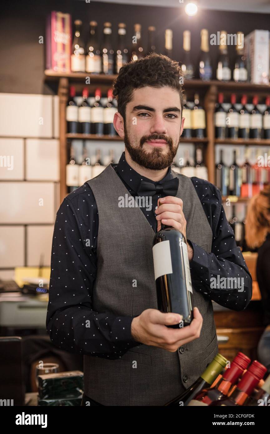 Homme adulte regardant l'appareil photo tout en tenant une bouteille de vin dans un magasin de vin. Banque D'Images