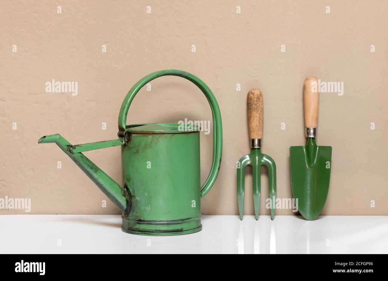 Ensemble de petits outils de jardin verts, dont une arrosoir à l'ancienne, une truelle et une fourche debout, alignés sur un mur beige Banque D'Images