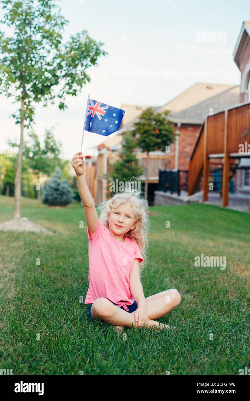 Bonne fille blonde de race blanche portant le drapeau australien. Enfant souriant assis sur l'herbe dans le parc et portant le drapeau de l'Australie. Enfant fêtant la fête de l'Australie Banque D'Images