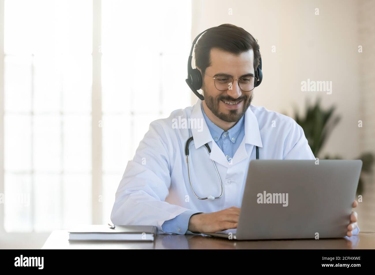 Un jeune médecin souriant dans un micro-casque/oreillette, consultant un patient en ligne, à l'aide d'un ordinateur Banque D'Images