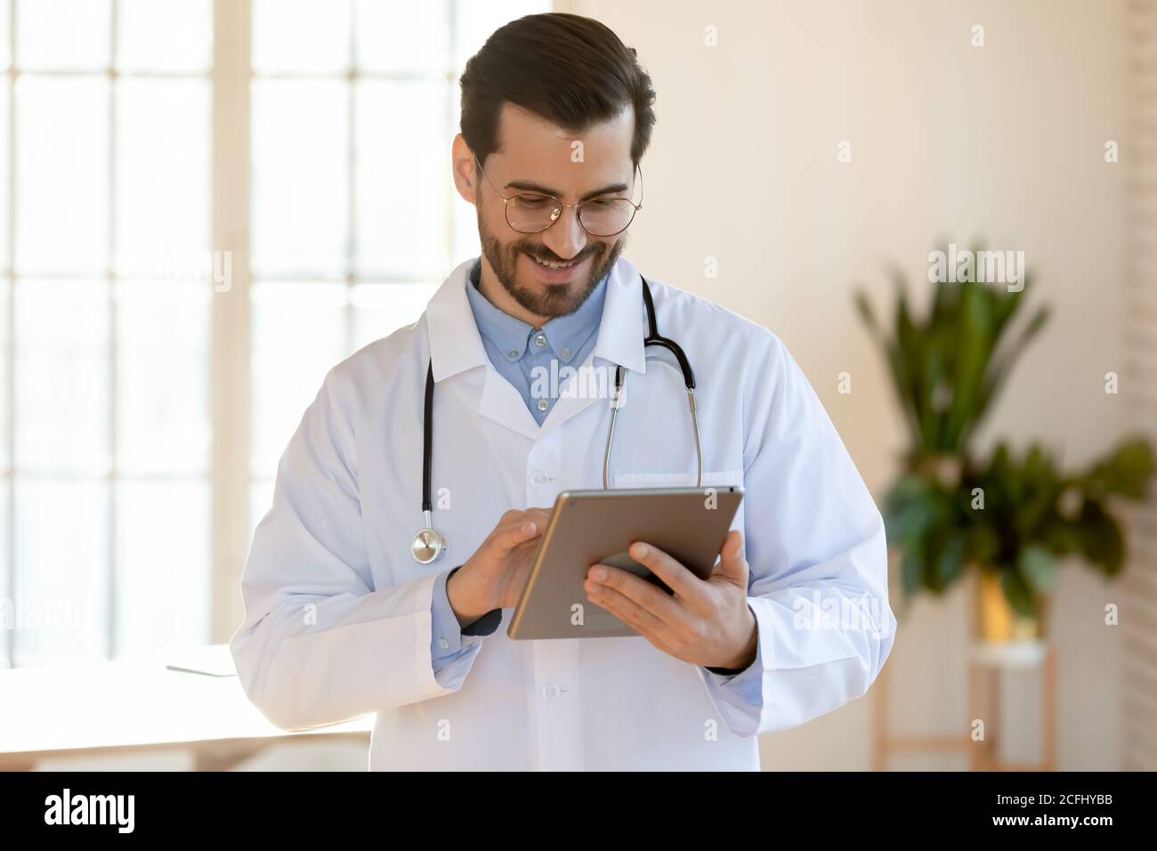 Médecin souriant portant des lunettes avec une tablette, debout au bureau Banque D'Images