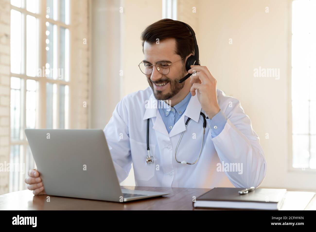 Un médecin souriant portant un micro-casque/une oreillette qui travaille sur un ordinateur portable, consulte le patient Banque D'Images