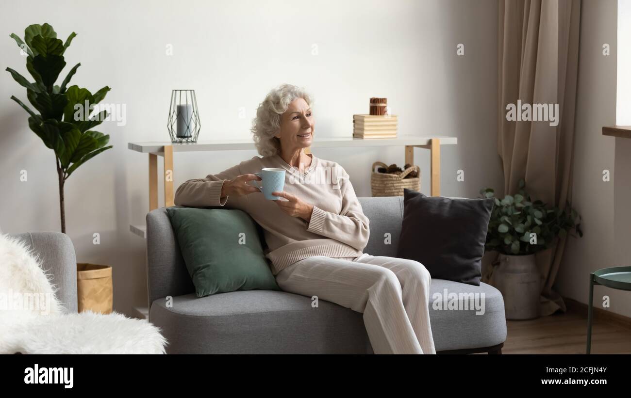 Une femme plus âgée souriante buvant du thé, assise sur un canapé confortable Banque D'Images