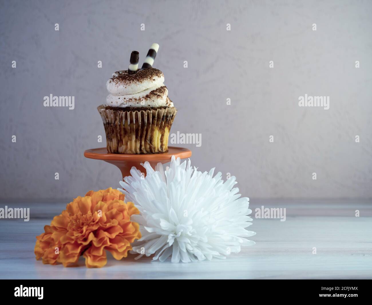 Tiramisu cupcake avec glaçage blanc arrosé de poudre d'espresso assis sur un support à cupcake orange avec un blanc et un orange fleur artificielle encore Banque D'Images