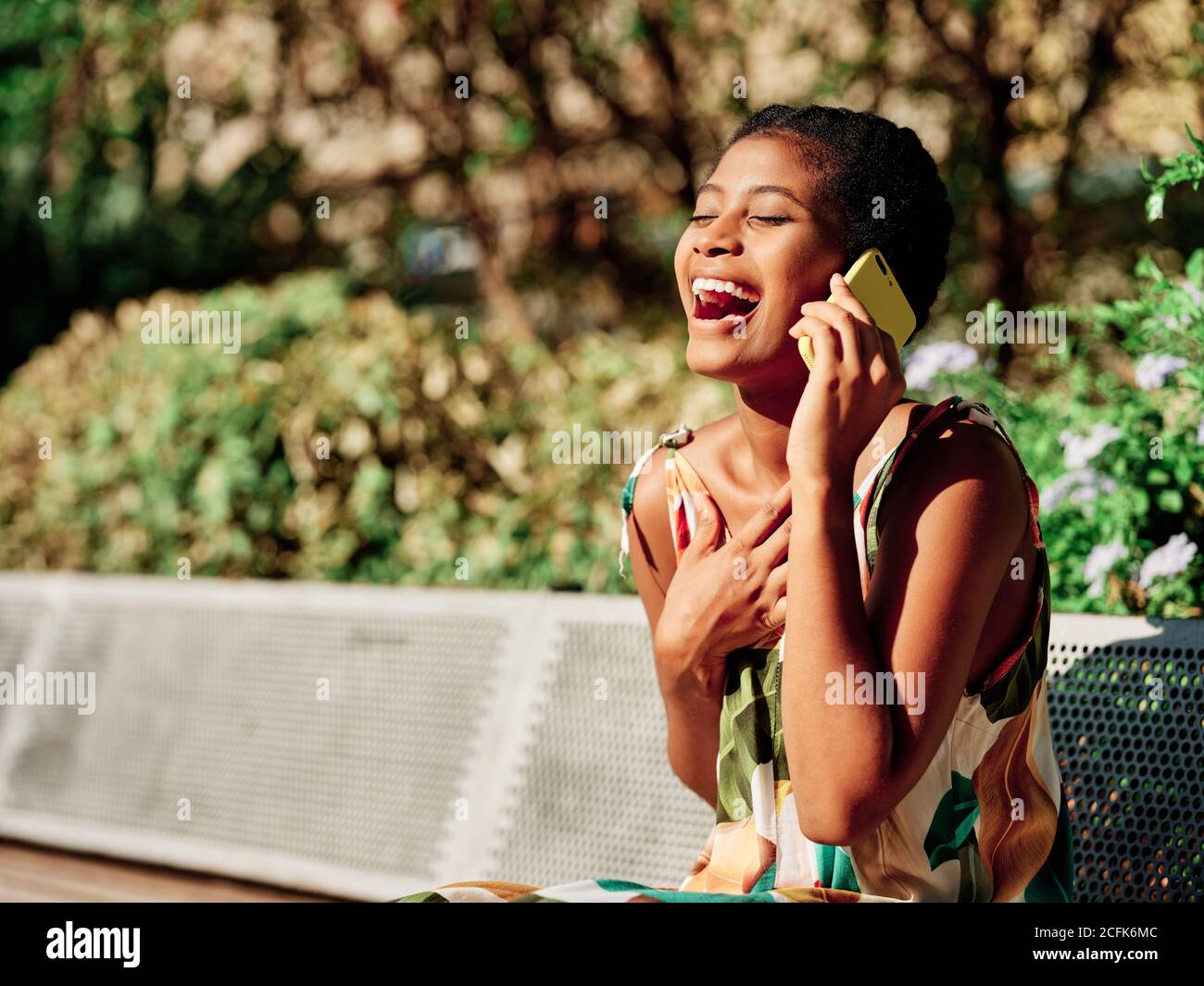 Jeune femme afro-américaine ravie dans une tenue élégante et colorée assise  sur le banc et discuter des nouvelles avec un ami pendant un appel  téléphonique tout en passant une journée d'été ensoleillée