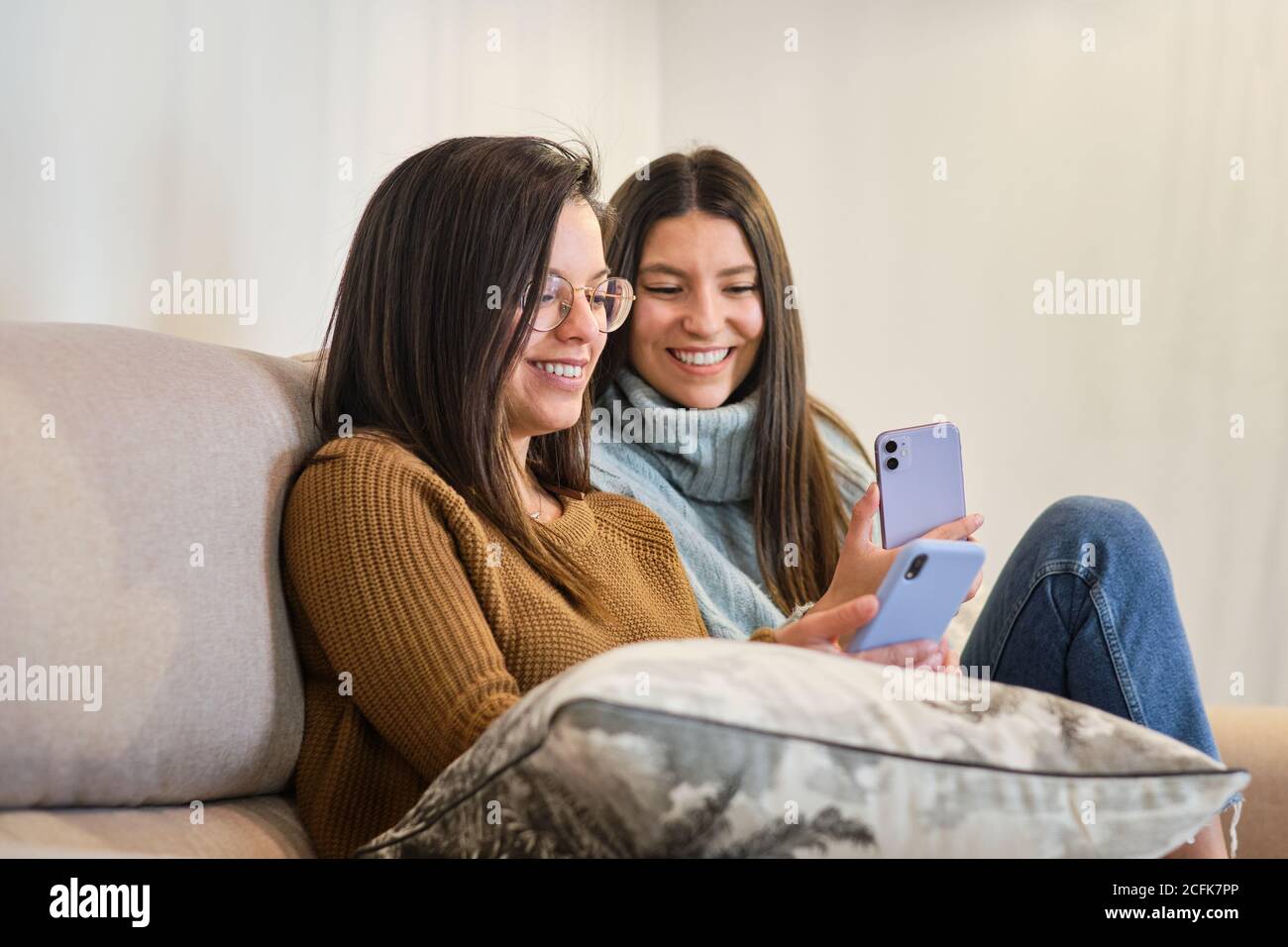 Des amies positives assises sur un canapé dans une chambre confortable et lire des messages sur les réseaux sociaux sur les téléphones mobiles tout en vous relaxant pendant le week-end à la maison Banque D'Images