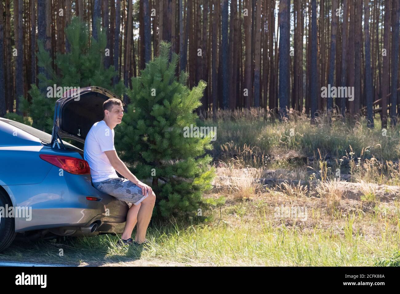Arrêter. Un jeune homme adulte s'assoit dans le tronc d'une voiture sur fond de forêt de pins le long de la route Banque D'Images