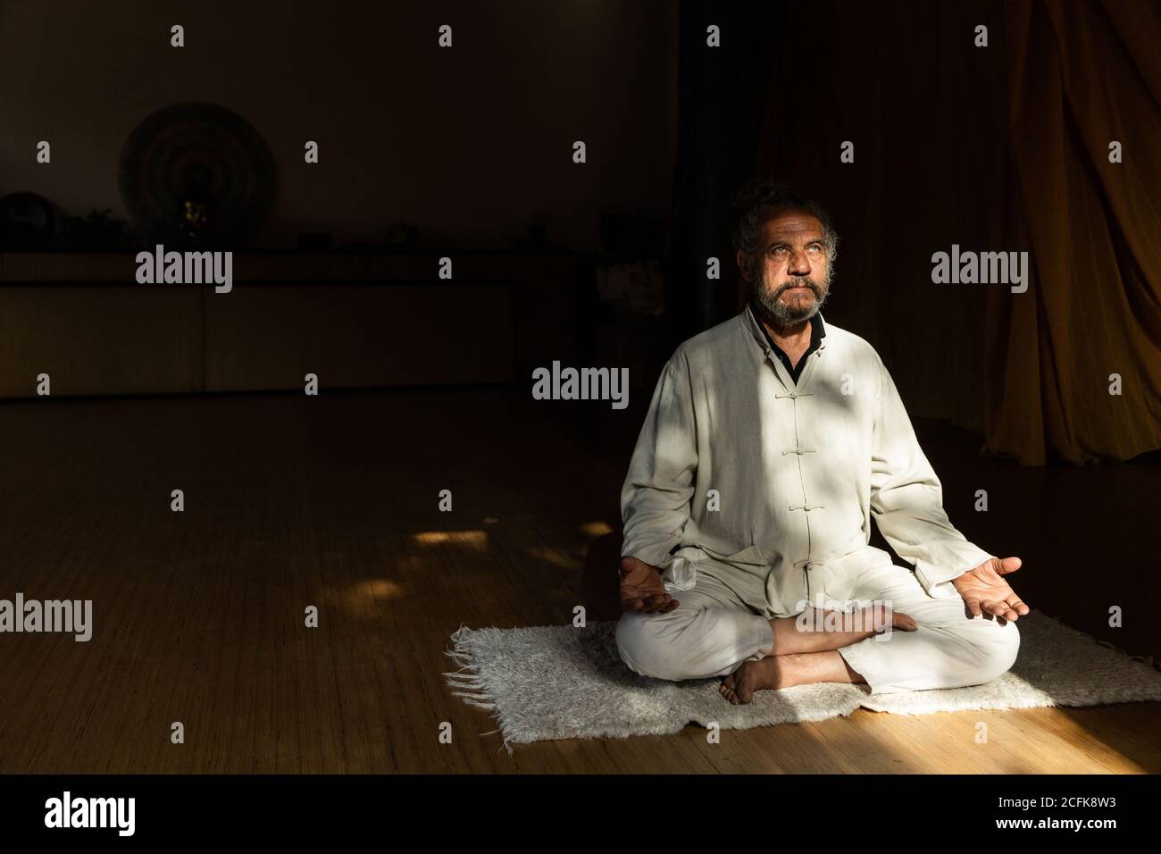 Vue latérale complète du corps d'un homme senior ethnique barbu pratiquer la méditation chi kung en position lotus dans un studio sombre Banque D'Images