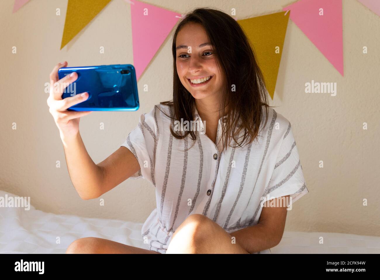 Vue latérale d'une femme enchantée assise sur un lit moelleux à accueil et lecture de messages sur les réseaux sociaux par téléphone mobile Banque D'Images