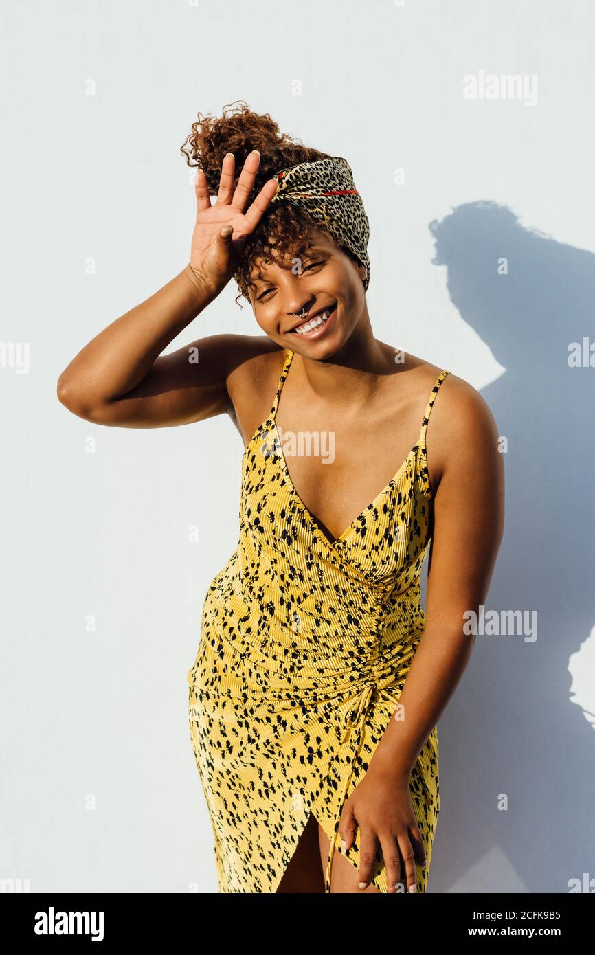Femme afro-américaine souriante portant une robe d'été et un bandeau  tendance se tenir près du mur en ville et toucher la tête tout en regardant  à l'appareil photo Photo Stock - Alamy
