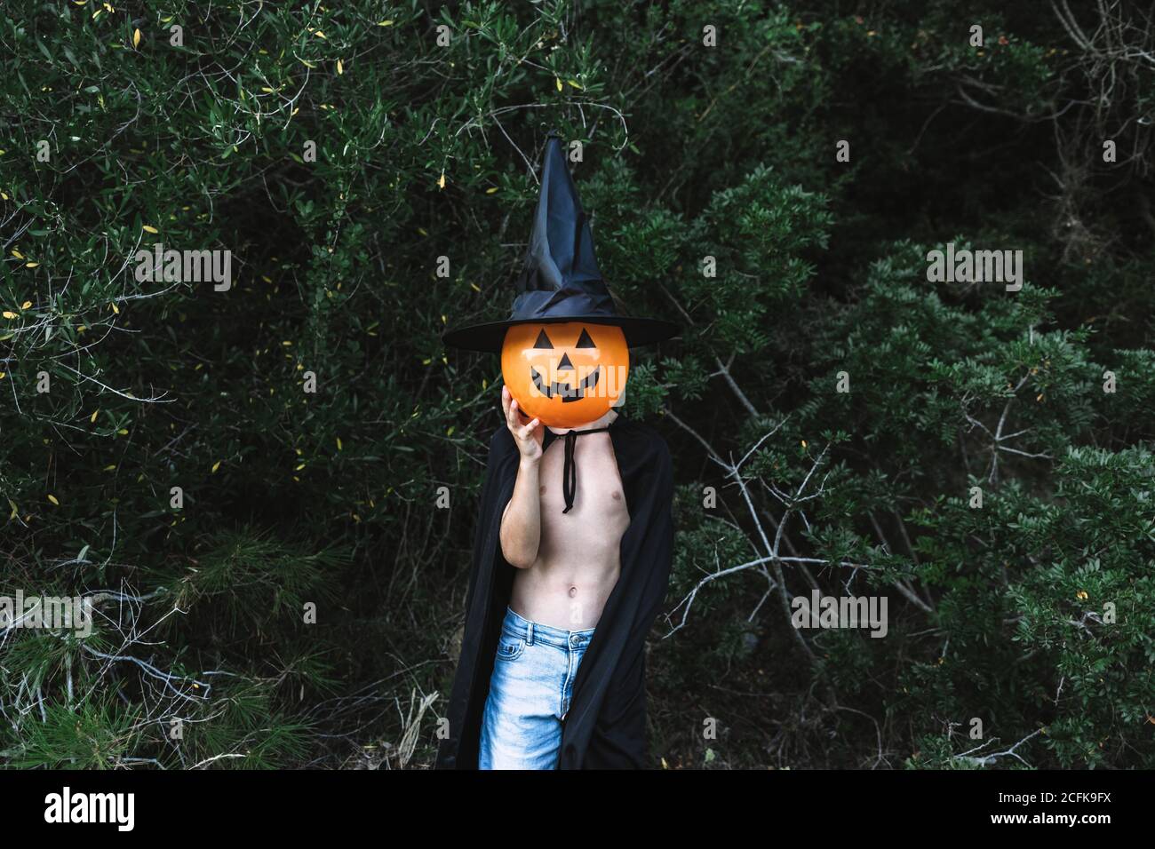 Enfant méconnaissable dans un manteau noir d'Halloween couvrant le visage avec de l'air ballon en forme de citrouille Banque D'Images