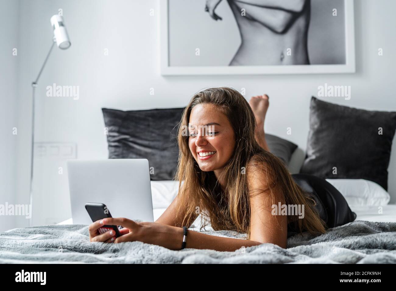 Positive femme en vêtement de nuit allongé sur un lit et une lecture doux messages sur le smartphone le matin après la veille Banque D'Images