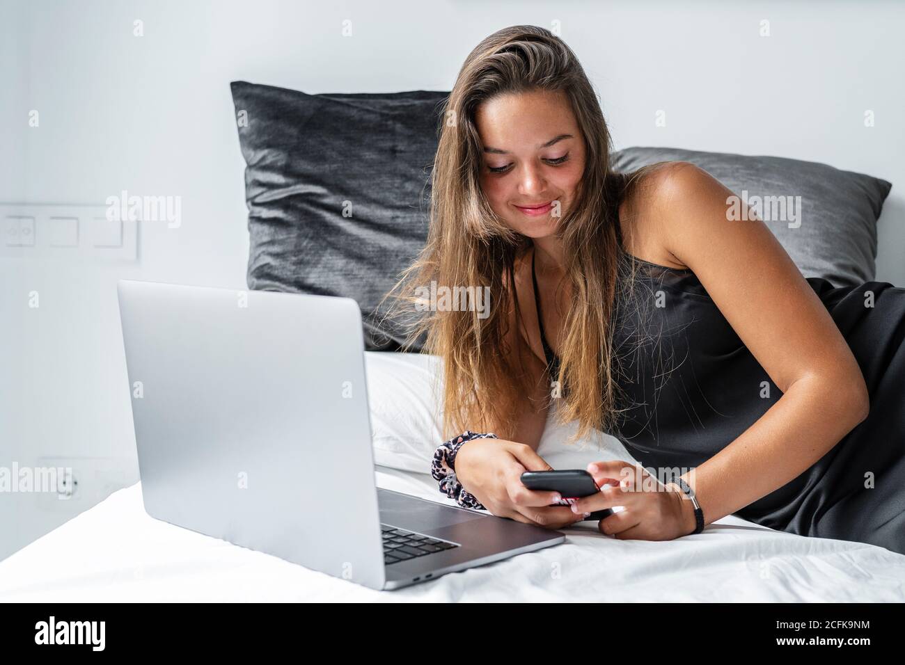 Positive femme en vêtement de nuit allongé sur un lit et une lecture doux messages sur le smartphone le matin après la veille Banque D'Images