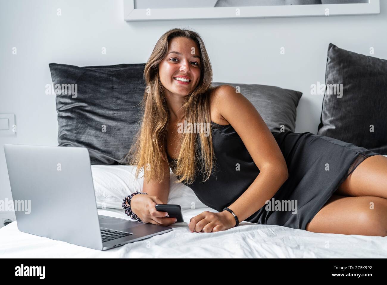 Positive femme en vêtement de nuit couché regardant l'appareil photo sur la douceur lit et lit les messages sur le smartphone le matin après le sommeil Banque D'Images