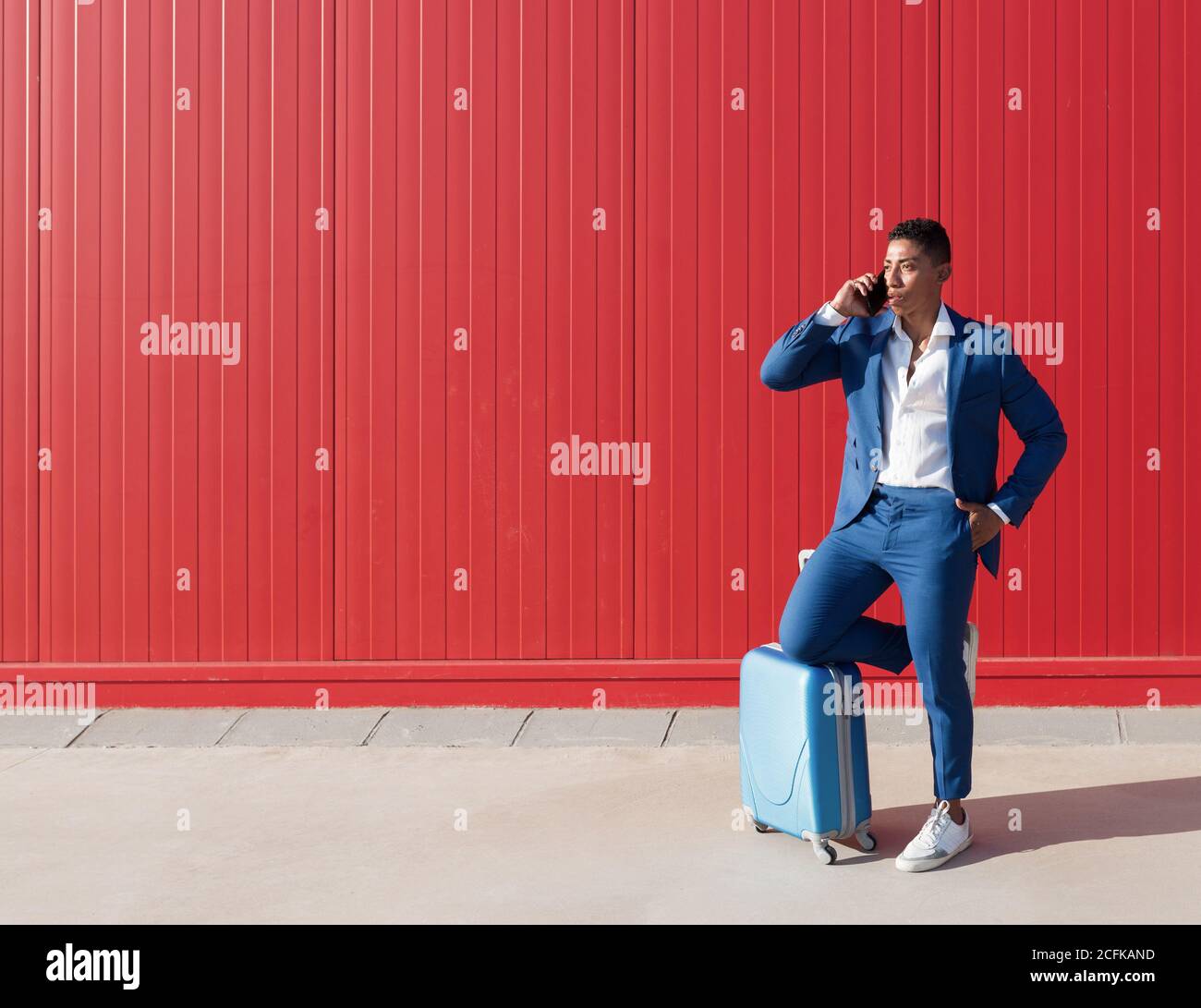 Corps complet du jeune homme afro-américain en bleu élégant combinaison avec messagerie bagages sur téléphone portable en vous tenant contre mur rouge sur la rue Banque D'Images