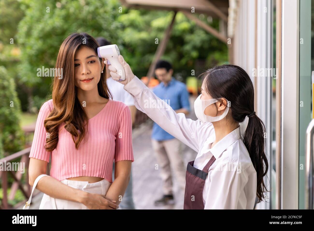 Serveuse asiatique avec masque facial prenant la température au client avant d'entrer dans le restaurant avec la file d'attente de distance sociale dans la file. Nouveau restaurant classique Banque D'Images
