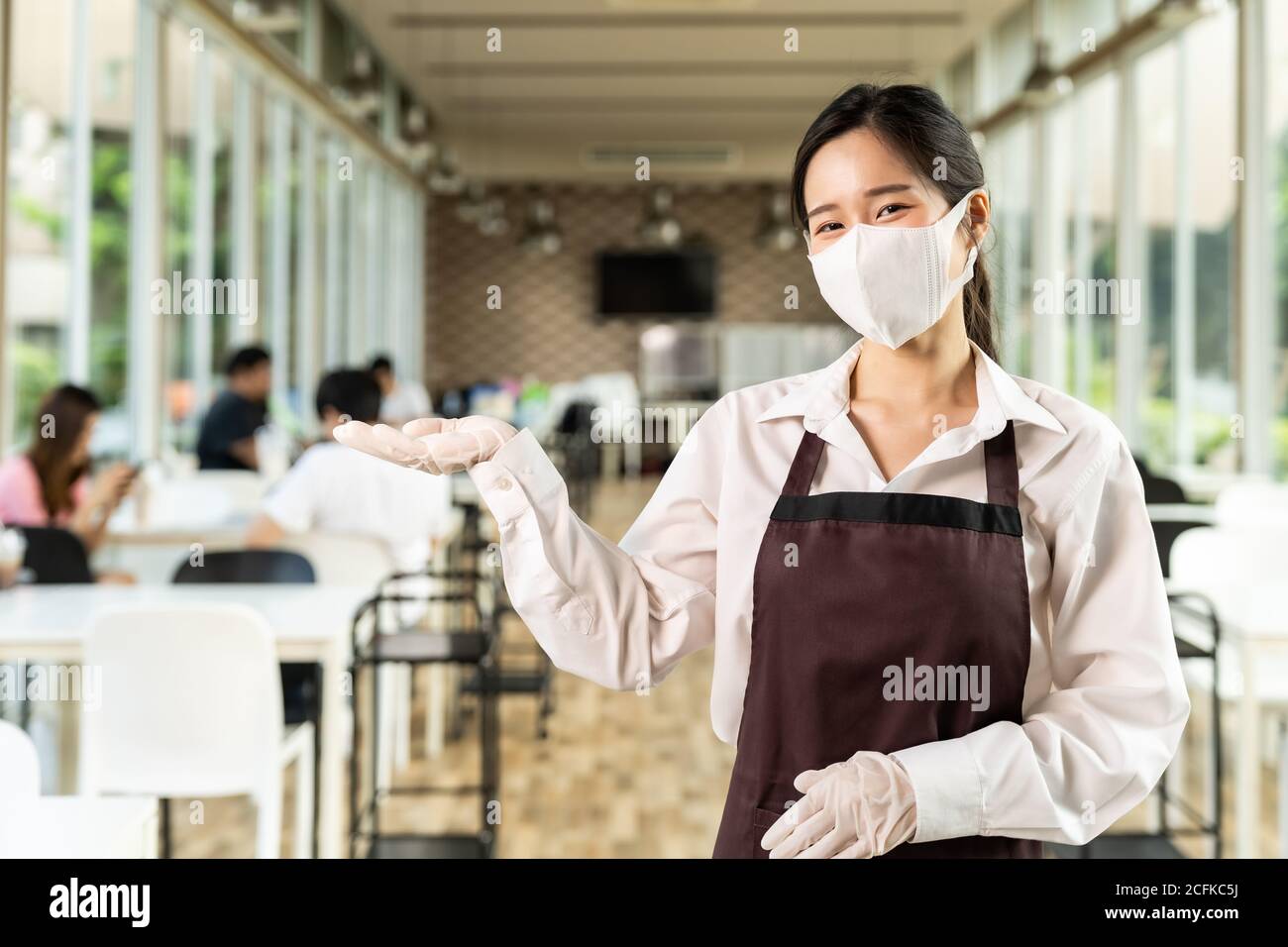 Portrait élégant asiatique porte-serveuse visage avec le fond des clients assis dans la table de social distance dans le restaurant à l'intérieur. Nouveau restaurant normal lif Banque D'Images
