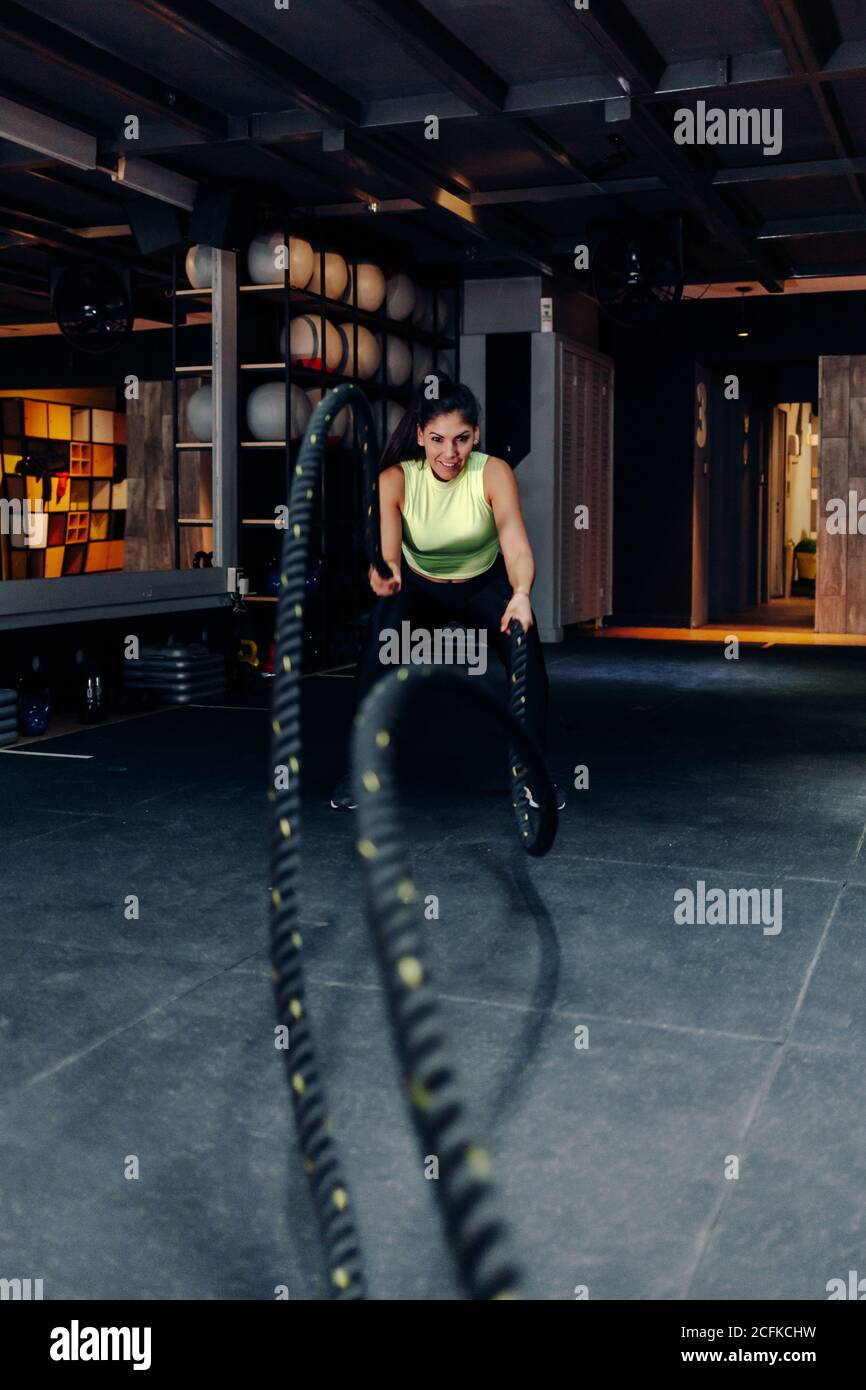 Jeune femme sportive déterminée et concentrée avec cordes de combat pendant séance d'entraînement intense dans la salle de sport moderne Banque D'Images