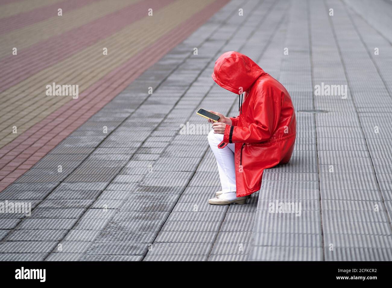 Une jeune femme en imperméable rouge a tiré un capot sur sa tête pour se cacher de la pluie. Assis sur les marches, fumer, regarder le smartphone Banque D'Images