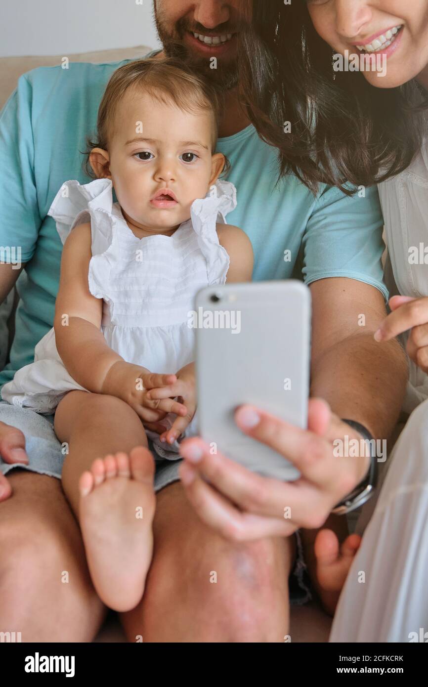 Photo verticale d'une petite fille dans une robe blanche et distrait l'expression sur les genoux de son père tout en faisant un selfie comme une famille à la maison Banque D'Images