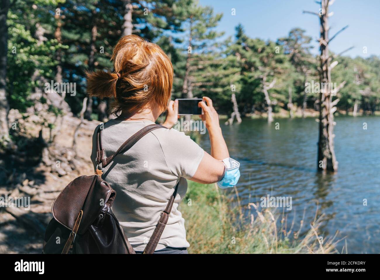 Femme mûre avec un masque médical prenant une photo avec un téléphone portable à un lac Banque D'Images