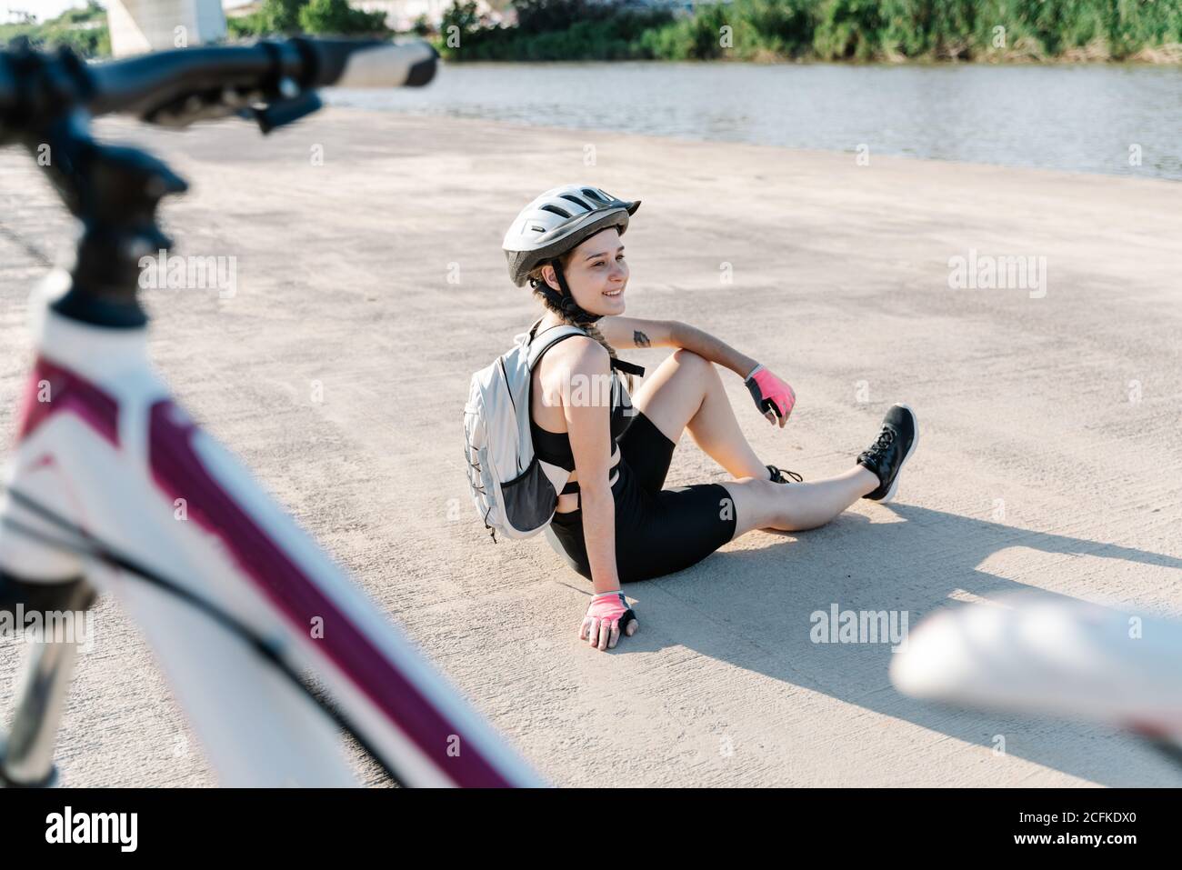 Vue latérale en grand angle de la jeune femme heureuse en vêtements de sport et casque avec sac à dos assis sur un sol pavé et reposant après avoir fait du vélo en été Banque D'Images