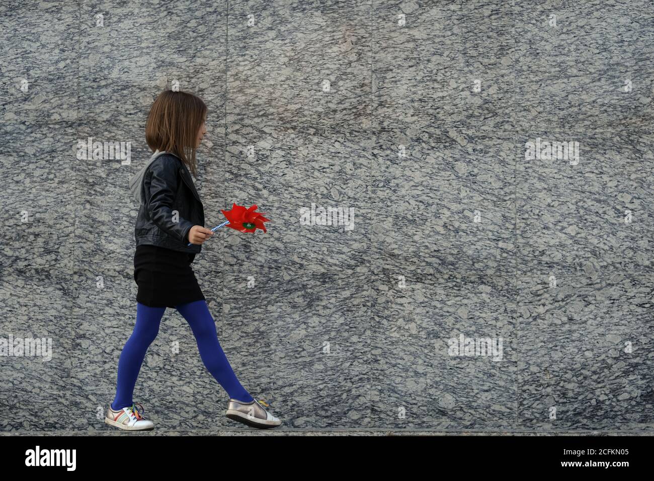 Petite fille urbaine élégante dans des vêtements d'automne décontractés marchant le long du mur de granit gris avec le vent rouge spinner. Espace pour le texte. Banque D'Images