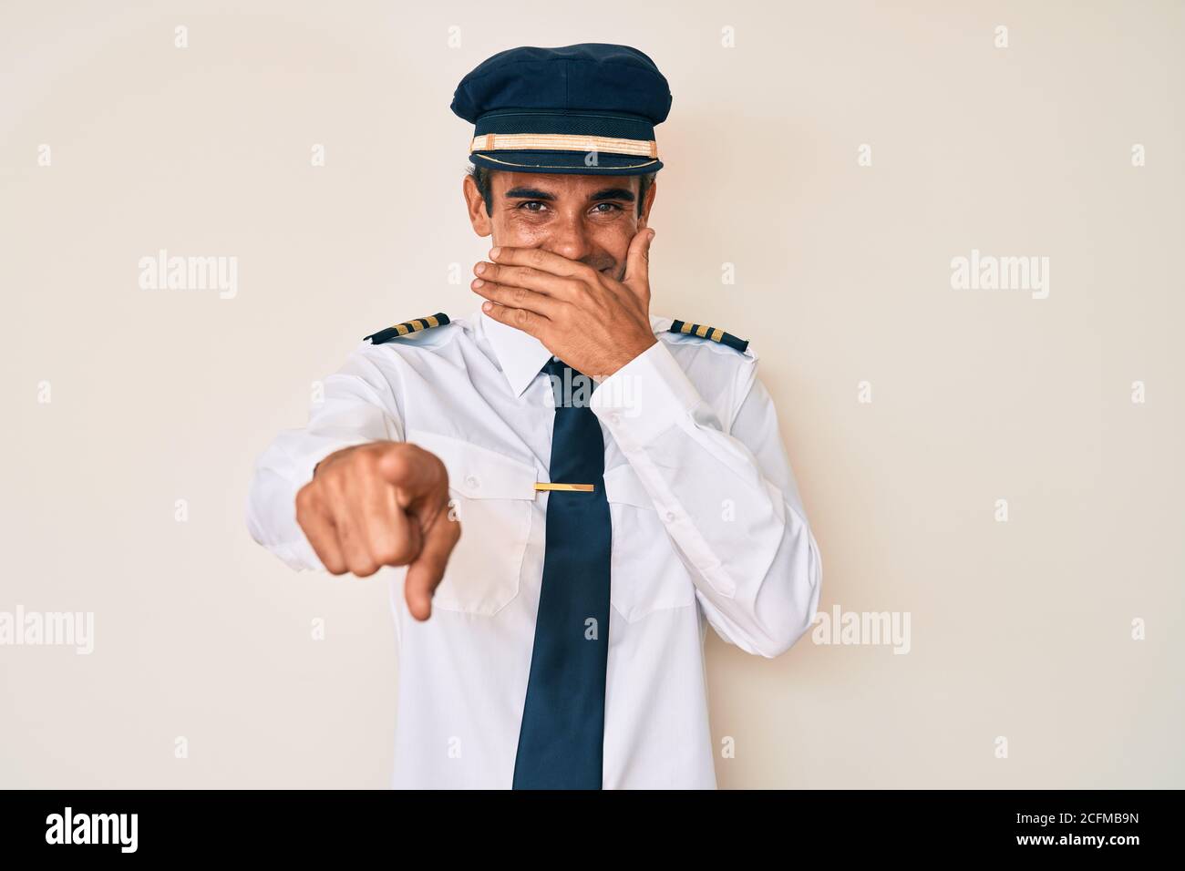 Jeune homme hispanique portant l'uniforme de pilote d'avion riant à vous, pointant le doigt vers la caméra avec la main sur la bouche, expression de honte Banque D'Images