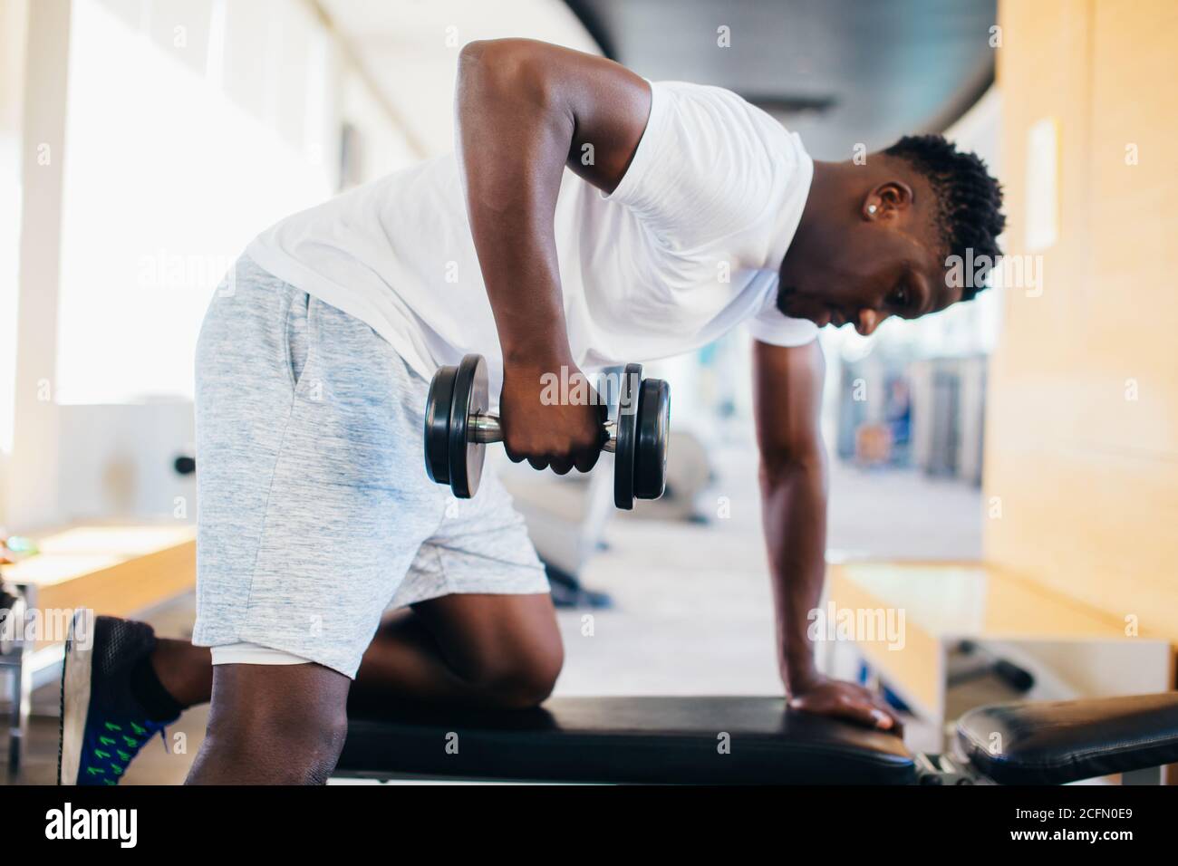 Vue latérale de l'homme africain américain dans les vêtements de sport se  penchant sur le banc et faire des exercices de rangée avec haltère pendant  l'entraînement dans la salle de gym Photo