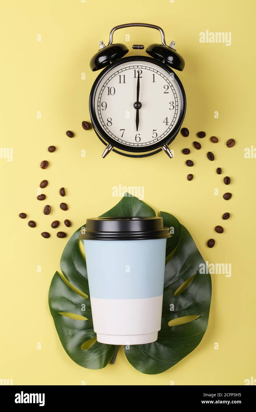 Tasse de café en papier sur une feuille de monstère et réveil sur fond jaune coloré. Concept de réveil du matin. Vue de dessus de la pose à plat Banque D'Images