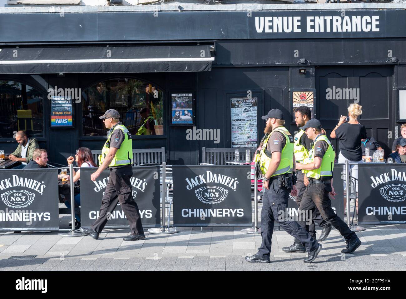 Une équipe de sécurité communautaire patrouilant sur le front de mer à Southend on Sea, Essex, Royaume-Uni, en passant devant un pub en bord de mer avec des buveurs à l'extérieur par beau temps Banque D'Images