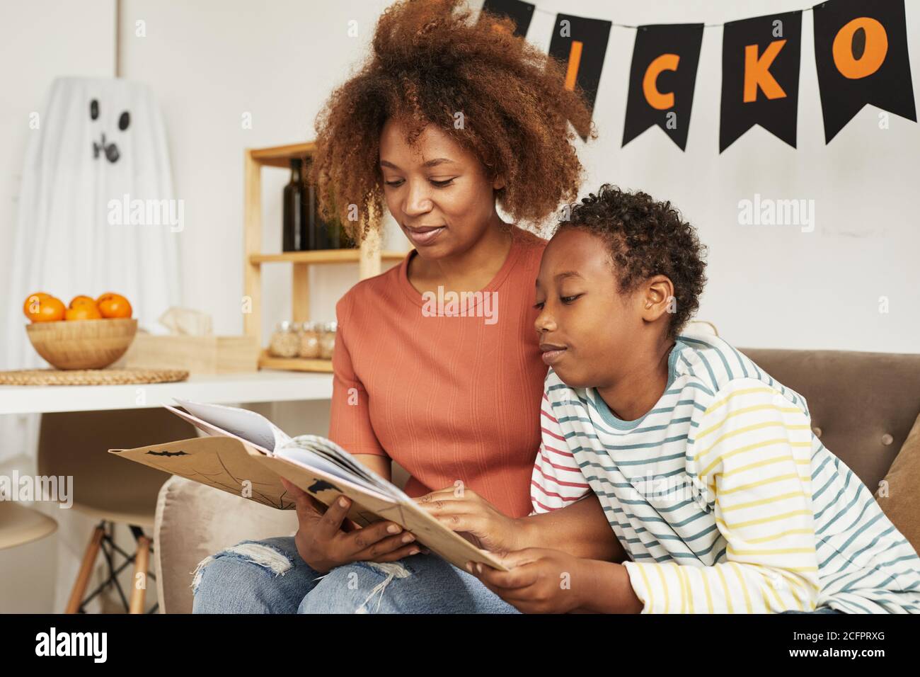 Belle femme afro-américaine assise sur un canapé dans la salle de séjour Avec son fils préadolescent lisant intéressant Halloween histoires livre Banque D'Images