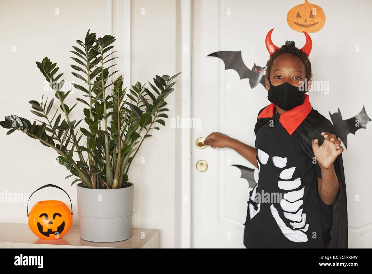 Un garçon américain africain moderne portant un costume de diable avec des cornes rouges et un masque noir sur le visage en disant au revoir à ses parents et en allant à trick ou à traiter avec des amis. Banque D'Images