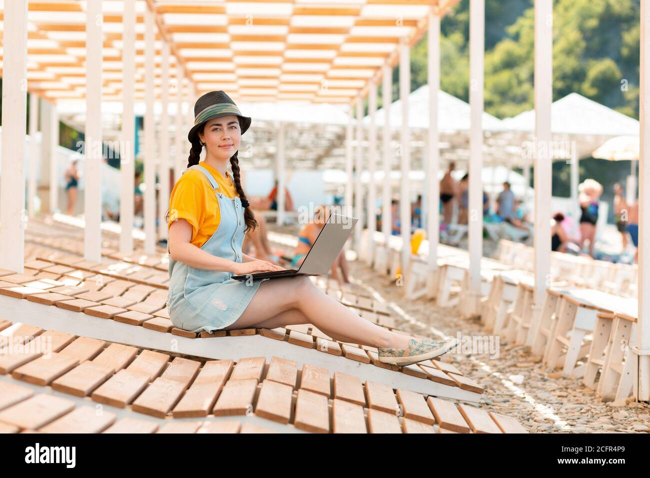 Une jeune jolie femme est assise sur une chaise longue avec un ordinateur portable et souriante. Parasols de plage en arrière-plan. Le concept de la pigiste et de la distance Banque D'Images