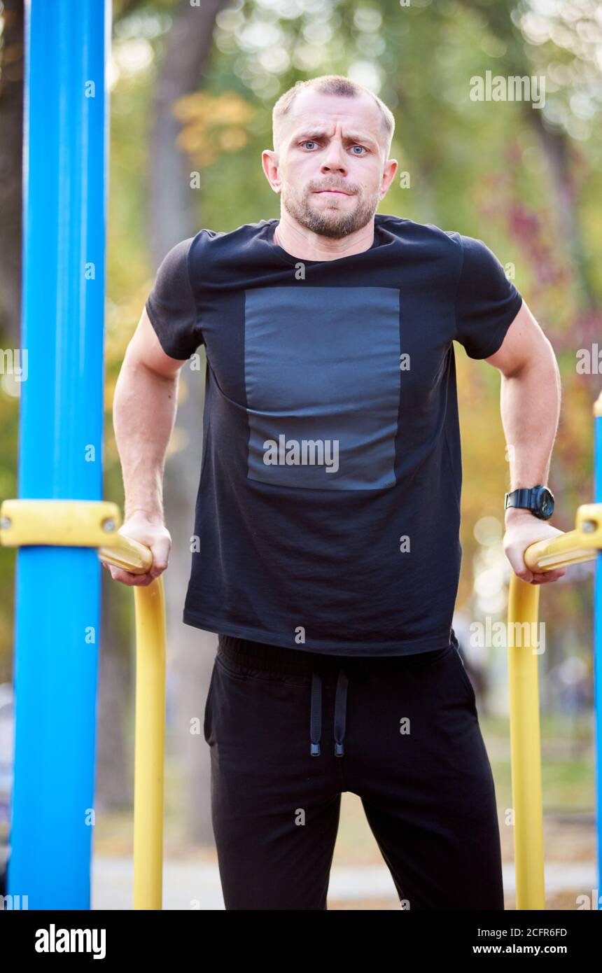 Portrait sportif homme vêtu d'une tenue noire est en train de faire de l'activité sur les barres parallèles à l'extérieur, concentré dans la vue de l'appareil photo, gros plan Banque D'Images