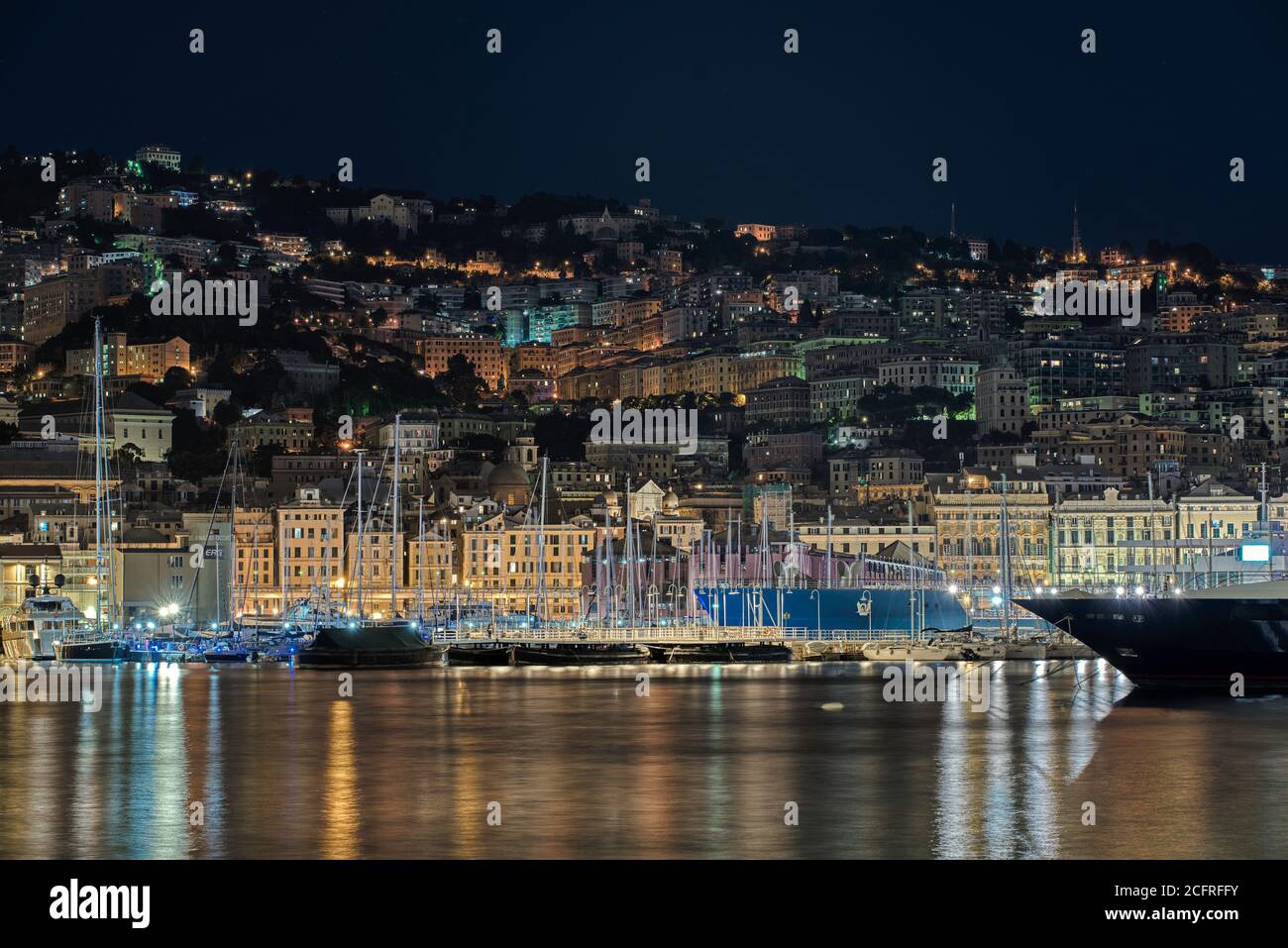 Grand bateau amarré dans le Vieux Port de Gênes, Italie la nuit devant des bâtiments illuminés de bord de mer et des yachts amarrés dans la marina avec des réflexions Banque D'Images